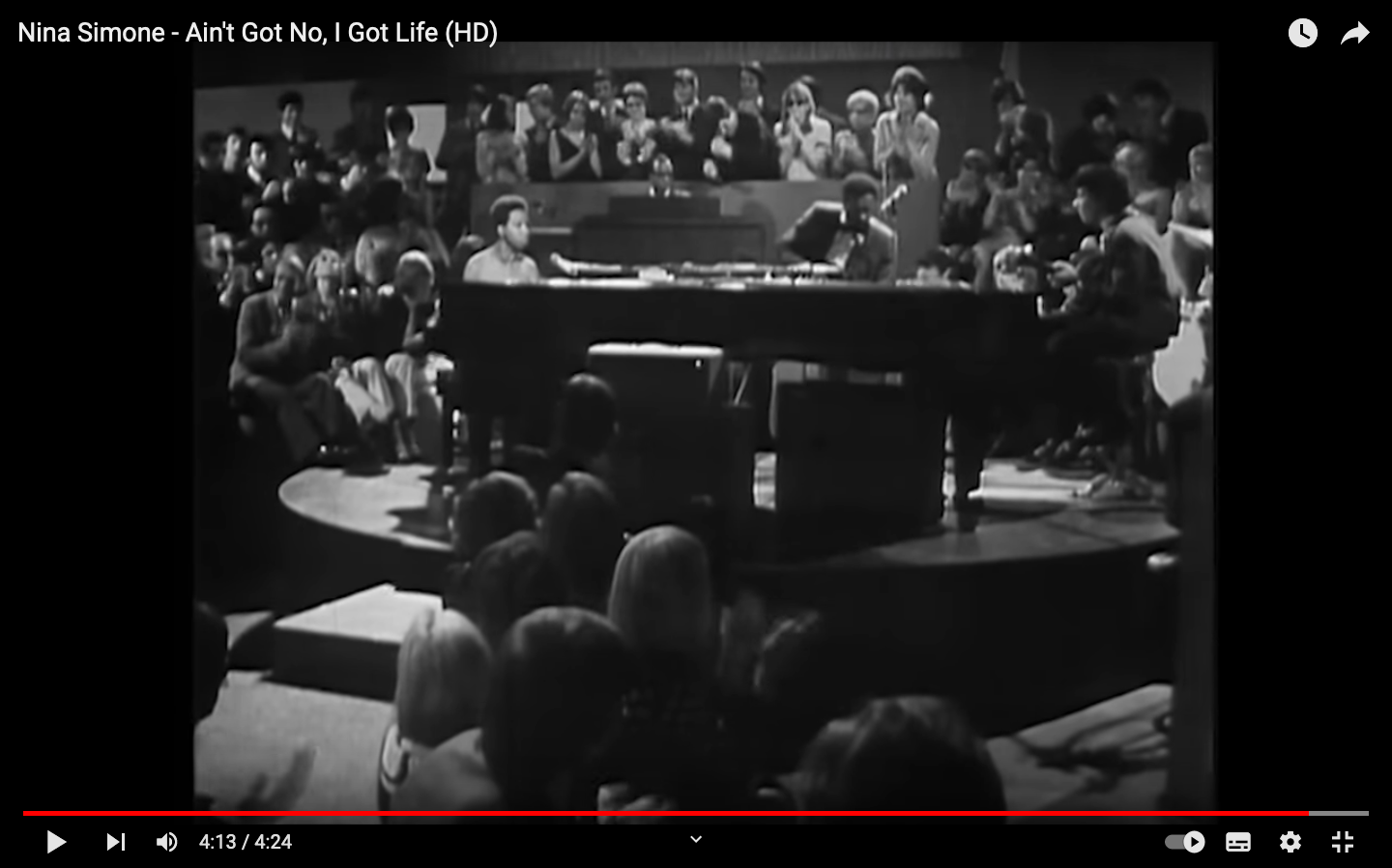 Capture d'écran YouTube de la vidéo du concert de Nina Simone. En noir et blanc. Au centre un énorme piano derrière Nina assise à celui-ci. Elle est entourée de ses musiciens. Le public est assis tout autour d'elle.